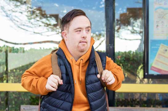 A man wearing a backpack and hoodie is standing at a bus stop
