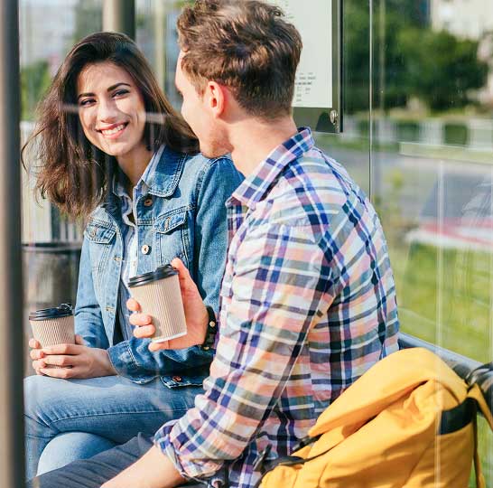 A man and a woman are sitting at a bus stop drinking coffee