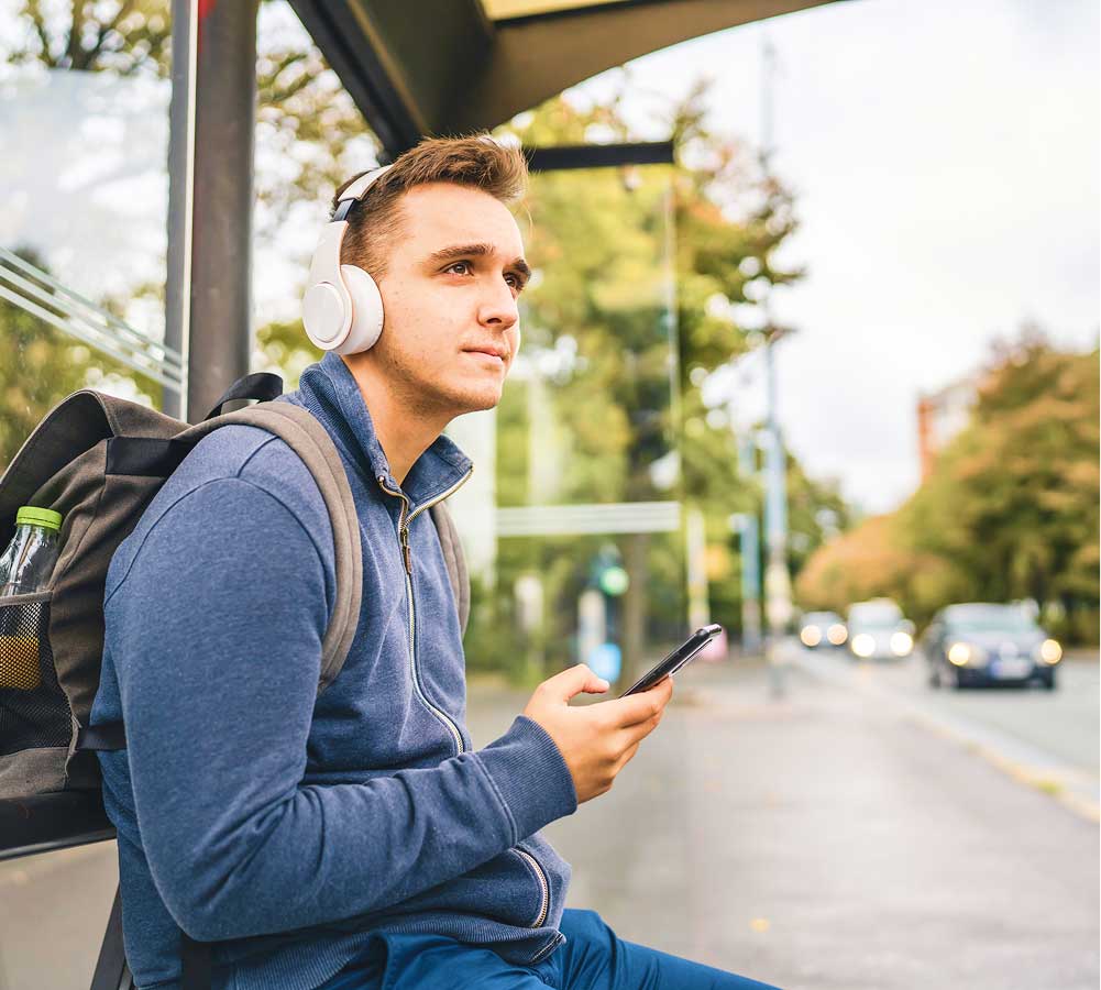 A man wearing headphones sits at a bus stop looking at his phone